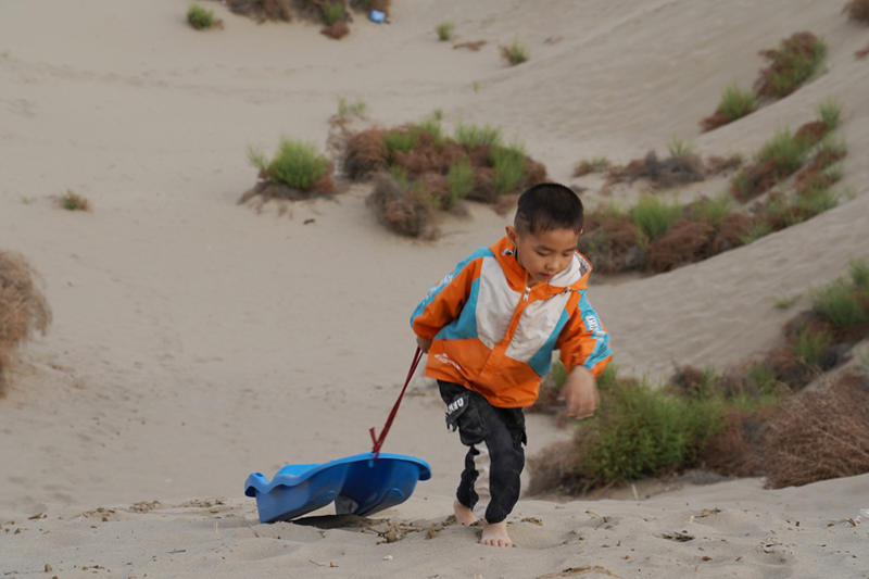 Preparándose para deslizarse por una pendiente de arena, un ni?o arrastra su trineo hasta la cima de la duna en la sección suroeste del desierto de Taklamakan en la región autónoma Uygur de Xinjiang, noroeste de China, el 21 de mayo de 2021 (Pueblo en Línea / Zhang Ruohan)